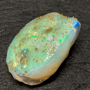 May include: A rough opal gemstone with a mix of colours. The opal has a base of white and blue, with patches of green and gold. The surface has a rough texture.
