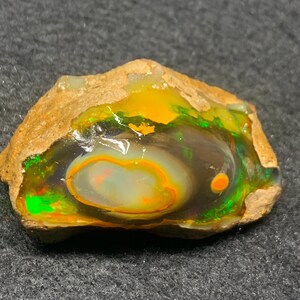 Puede incluir: Una piedra de ópalo en bruto con una vibrante exhibición de colores. El ópalo presenta un remolino central de blanco y naranja, rodeado de bandas de naranja, verde y negro. La capa exterior es una matriz de color beige.