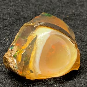 Puede incluir: Una piedra preciosa de ópalo en bruto con un centro translúcido y redondeado. El ópalo muestra una mezcla de colores, incluyendo naranja, amarillo y toques de verde y marrón. La piedra está sobre un fondo oscuro y texturizado.