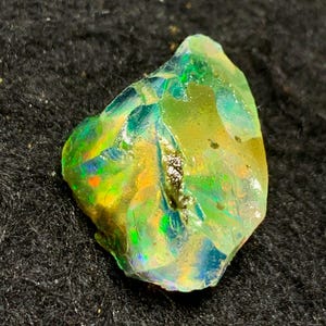 Könnte beinhalten: Ein lebendiger, roher Opal-Edelstein mit einer Mischung aus Farben, darunter Grün, Gelb, Blau und Orange. Der Opal hat eine raue, unregelmäßige Form und zeigt ein Farbenspiel. Der Edelstein liegt vor einem dunklen, strukturierten Hintergrund.