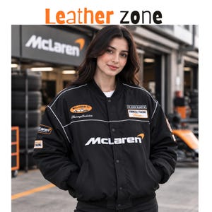 Op de afbeelding: Een zwart bomberjack met het McLaren-logo in wit en oranje, voorzien van diverse race-gerelateerde patches op de borst en mouwen. Het jack heeft een sluiting met drukknopen, geribbelde boorden en witte biezen over de schouders.