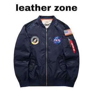 Puede incluir: Chaqueta bomber azul marino con parches de la NASA y la bandera estadounidense. Cuenta con cierre de cremallera, dos bolsillos delanteros y una etiqueta roja "REMOVE BEFORE FLIGHT". El texto "leather zone" está en la parte superior.