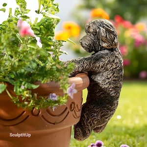 May include: A bronze-colored dog sculpture clinging to the edge of a terracotta flower pot filled with vibrant flowers. The dog has a textured coat and is positioned as if it's peering into the pot. The scene is set outdoors with a blurred background of colorful blooms.