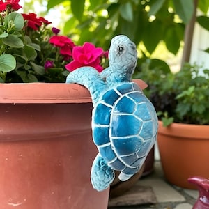 May include: A blue turtle-shaped planter decoration. The turtle has a detailed shell pattern and is perched on the edge of a terracotta-colored flower pot. Red and pink flowers are visible in the background, with green foliage.