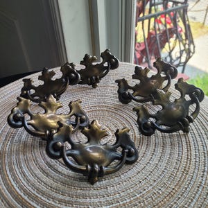 以下が含まれることがあります： 真鍮製のアンティーク引き出し取っ手6個セット。各取っ手は装飾的な葉のデザインと湾曲したハンドルが特徴です。それらは、背景に窓がある、質感のある表面に配置されています。