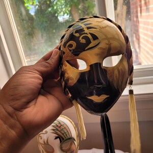 Pode incluir: Uma máscara decorativa preta e dourada com uma borda trançada e borlas creme. A máscara apresenta um design pintado com detalhes dourados e é segurada em frente a uma janela.