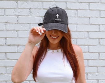 May include: A dark gray baseball cap with the text "I Believe" and a white cross. The cap has a curved brim and an adjustable strap. The background is a white brick wall.