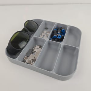 May include: A gray, divided organizer tray filled with various small items. The tray contains a pair of black sunglasses, small silver discs, and several black and blue fittings. The tray is on a white surface.