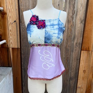May include: A sleeveless top with a blue and white tie-dye bodice, adorned with two magenta fabric flowers. The waist features a decorative band and a lavender skirt with a white script design and a lace trim. The top is displayed on a mannequin.