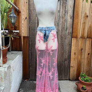 May include: A mannequin displays a unique garment. It features a pair of denim shorts with a sheer, pink, floor-length skirt overlay. The skirt is adorned with intricate floral lace detailing. The ensemble is set against a wooden fence backdrop.