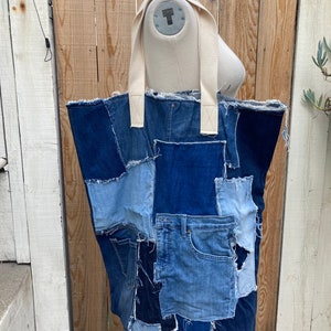 May include: A large tote bag made from repurposed denim fabric. The bag has a patchwork design with different shades of blue denim. The bag has two white canvas straps.