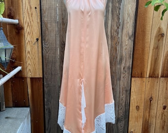 May include: A peach-colored satin halter dress with a white lace hem. The dress is displayed on a mannequin. The dress has a high neckline and a flowing, asymmetrical hemline. The white lace trim adds a delicate contrast to the peach fabric.