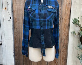 May include: A blue and black plaid long-sleeved shirt with a black denim bodice. The shirt has a button-up front and two chest pockets.