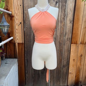 May include: A peach-coloured halter crop top featuring a high neckline adorned with a white lace trim. The garment is sleeveless, fitted through the torso, and includes a long fabric tie extending from the bottom hem, displayed on a mannequin.