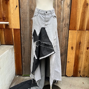 May include: A gray denim skirt with a distressed design, featuring a black fabric panel. The skirt has a high waist and a long, asymmetrical hemline. The skirt is displayed on a mannequin against a wooden background.
