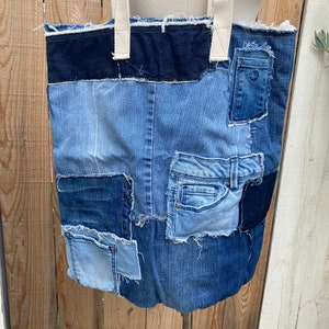 May include: A large blue denim tote bag made from repurposed jeans. The bag has a patchwork design with different shades of blue denim and a black denim trim. The bag has a white canvas strap.