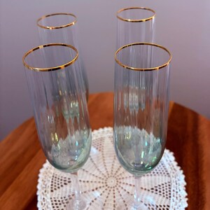 May include: Four clear champagne flutes with gold rims. The flutes are arranged on a white lace doily, with a brown surface in the background. The glasses have a vertical ribbed design.