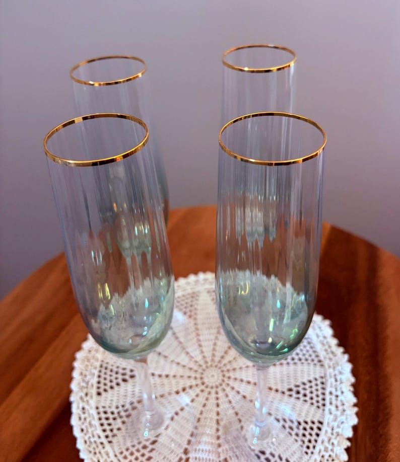 May include: Four clear champagne flutes with gold rims. The flutes are arranged on a white lace doily, with a brown surface in the background. The glasses have a vertical ribbed design.