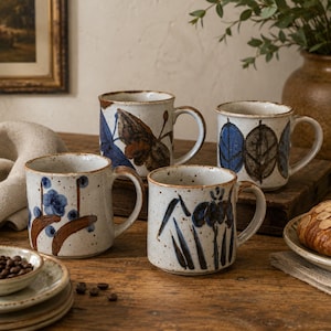 May include: Four ceramic mugs with a speckled off-white glaze and hand-painted botanical motifs in shades of blue and brown. Each mug features a sturdy handle and a cylindrical shape, showcasing unique leaf, floral, and stem patterns.