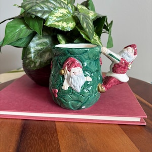 May include: A ceramic Christmas mug with a green holly design and Santa Claus figures. The handle is a Santa Claus figure. The mug is sitting on a red book, with a green plant in the background.