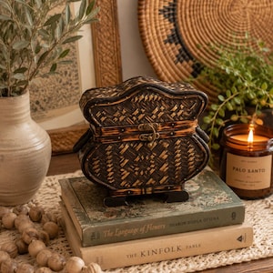 May include: A dark brown woven bamboo box with a curved lid, a metal latch, and four small feet. The box is resting on two stacked books titled The Language of Flowers and The Kinfolk Table, featuring a textured weave and burnt wood accents.