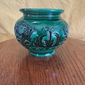 May include: A green ceramic vase with a glossy finish. The vase features a rounded body with a flared rim and base. Darker green and black floral patterns adorn the exterior. The vase sits on a wooden table.