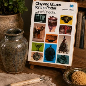 May include: A hardcover book titled Clay and Glazes for the Potter by Daniel Rhodes, featuring a grid of various ceramic pottery images on the cover. The book is displayed on a small wooden stand next to a ceramic vase and a pottery tool.