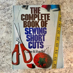 May include: A vintage book titled "The Complete Book of Sewing Short Cuts" by Claire B. Shaeffer. The cover features a pair of red scissors, a tomato pin cushion, and a yellow measuring tape.