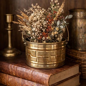 May include: A brass bucket with a geometric design holds a dried flower arrangement. The bucket sits atop two antique leather-bound books on a wooden shelf. A brass candlestick and a decorative container are in the background.
