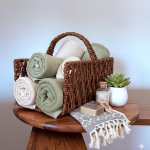 May include: A woven brown basket filled with rolled green and cream-colored towels. A small white pot with a succulent, a bar of soap, a small bottle with a cork, and a patterned towel with fringe are on a wooden table.