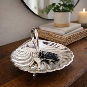 May include: A silver-colored, shell-shaped decorative tray with a handle, holding keys and a car remote. The tray sits on a dark wooden surface, with a mirror, plant, books, and candle in the background.