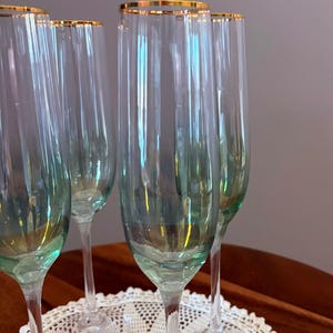 May include: A set of six champagne flutes with a gold rim. The clear glass flutes have a slight green tint and are displayed on a white lace doily. The stems are clear and slender.