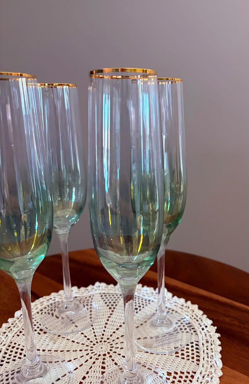 May include: A set of six champagne flutes with a gold rim. The clear glass flutes have a slight green tint and are displayed on a white lace doily. The stems are clear and slender.