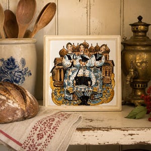 May include: A square ceramic tile featuring a colorful illustration of three individuals in traditional folk attire singing while one plays an accordion, set against a backdrop of onion-domed buildings, framed by a simple off-white border.