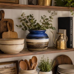 May include: A rustic shelf displays kitchenware and decor. Items include wooden cutting boards, ceramic bowls, a blue and brown striped vase with greenery, a brass watering can, and cookbooks. The shelf is made of dark wood.