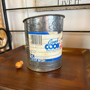 May include: A vintage 3-cup sifter made of metal with a wooden handle. The sifter has a label that reads "Good Cook" and "Quality by Design". The sifter is silver with blue and red accents. The sifter is sitting on a wooden tray.