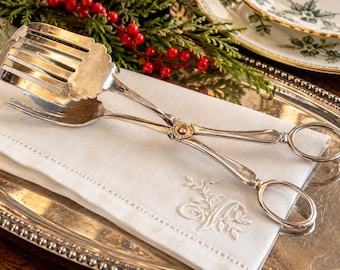 May include: A silver-plated serving tong featuring a slotted spade-shaped end and a matching fork end, connected by a central hinge mechanism with two circular finger loops for handling, resting on a white embroidered linen napkin.
