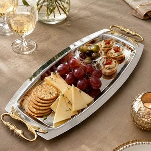 May include: A rectangular silver-toned serving tray with ornate gold-toned metal handles at each end. The tray holds a small glass bowl of green and black olives, a cluster of red grapes, sliced yellow cheese, and several round crackers.