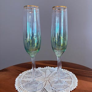 May include: Two iridescent champagne flutes with gold rims sit on a white lace doily. The flutes have a clear stem and a pearlescent finish that reflects shades of green and blue. The doily is placed on a dark wooden table.
