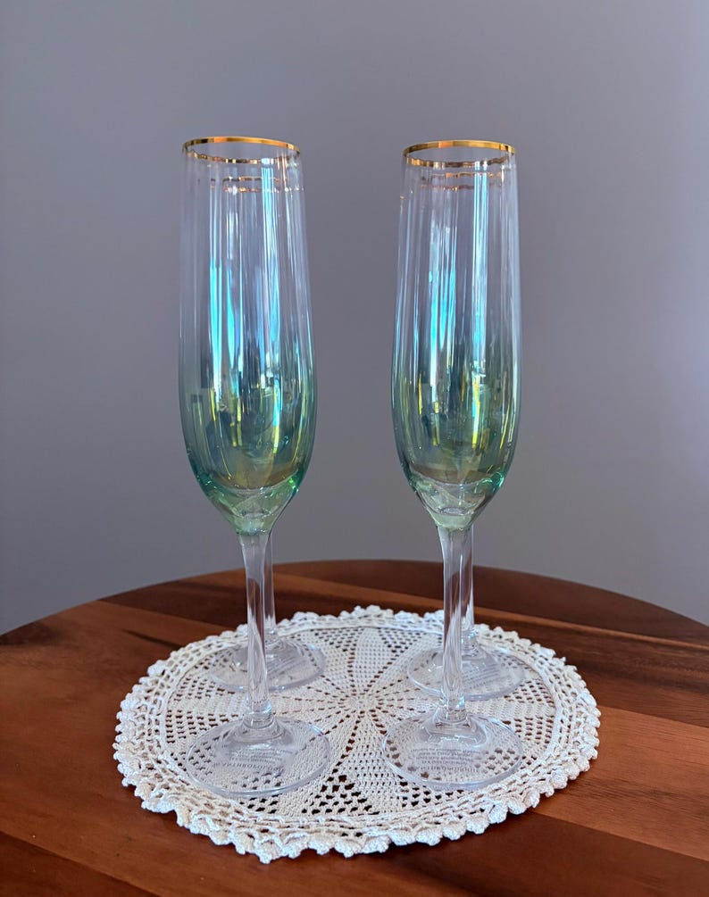 May include: Two iridescent champagne flutes with gold rims sit on a white lace doily. The flutes have a clear stem and a pearlescent finish that reflects shades of green and blue. The doily is placed on a dark wooden table.