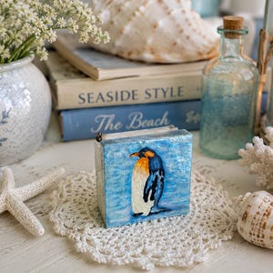 May include: A small, square, blue box with a hand-painted penguin illustration. The penguin has orange, black, and white markings. The box is on a white lace doily, with seashells, books, and a vase of flowers in the background. The books have the titles "Seaside Style" and "The Beach".