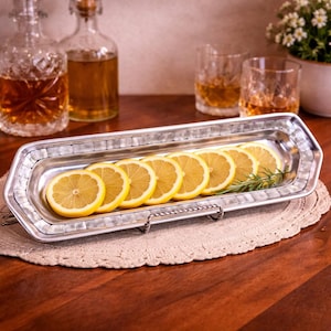 May include: A rectangular silver serving tray with a mother-of-pearl border holds sliced lemons and a sprig of rosemary. The tray sits on a beige doily, with bottles and glasses in the background. The tray is supported by a silver metal stand.
