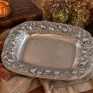 May include: A rectangular silver-toned serving tray with a decorative border of leaves and acorns. The tray has a slightly raised rim and a smooth, reflective surface. The tray is likely made of metal and is suitable for serving food or displaying decorative items.
