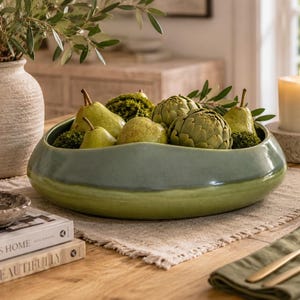 May include: A low, wide ceramic bowl with a smooth, two-toned green glaze. The bowl is filled with several green pears, artichokes, and small mossy spheres. The exterior features a darker sage green upper rim and a lighter lime green base.