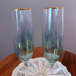 May include: Two champagne flutes with gold rims. The clear glass has a subtle blue-green tint. The flutes are on a white lace doily, with a wooden table in the background. The flutes are elegant and suitable for special occasions.