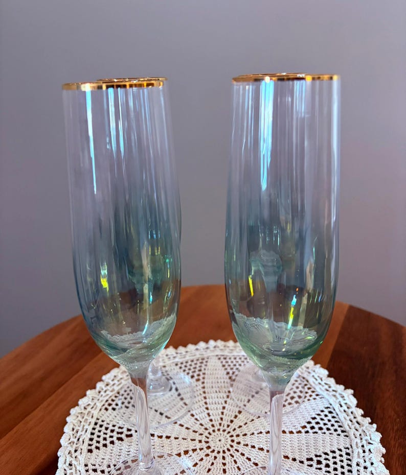 May include: Two champagne flutes with gold rims. The clear glass has a subtle blue-green tint. The flutes are on a white lace doily, with a wooden table in the background. The flutes are elegant and suitable for special occasions.