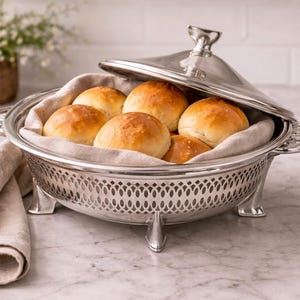 May include: A silver bread warmer with a lid, containing several golden-brown rolls. The warmer has ornate handles and rests on three feet. A linen napkin is draped nearby. A basket of greenery is in the background.