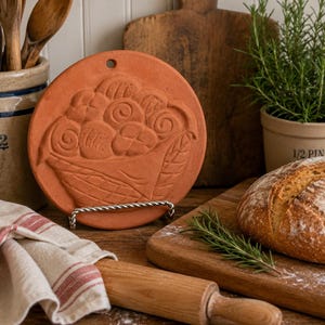 May include: A round, terracotta-colored clay baking mold featuring a relief design of a basket filled with various bread rolls. The mold has a small hole at the top for hanging and is supported by a silver-toned metal wire stand.