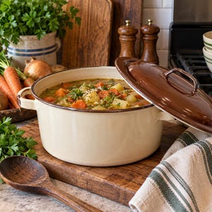 May include: A round, cream-colored enameled metal pot filled with vegetable soup, featuring a matching brown lid resting partially open on the rim. The pot has two side handles and a smooth finish, showcasing a hearty stew with visible carrots and potatoes.