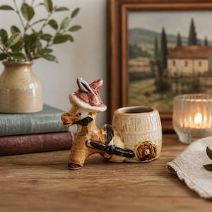 May include: A ceramic donkey figurine with a barrel on its back, wearing a red hat and black harness. The donkey is tan and white, with a small opening on the barrel. A small vase with greenery is in the background.